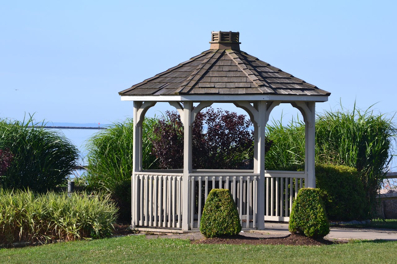 A charming wooden gazebo surrounded by lush greenery near a calm waterfront.