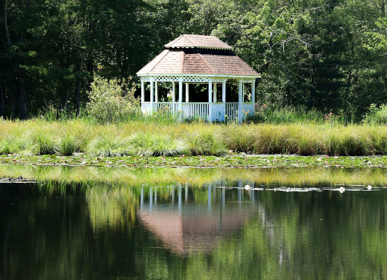 Charming white gazebo reflected in a peaceful pond, surrounded by lush greenery and trees.