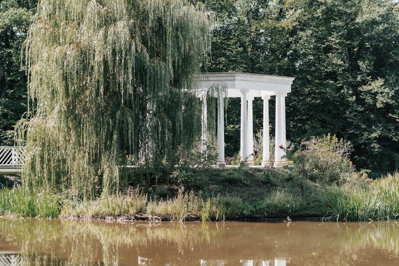 A peaceful garden scene with a classic white gazebo by a calm pond surrounded by lush greenery.
